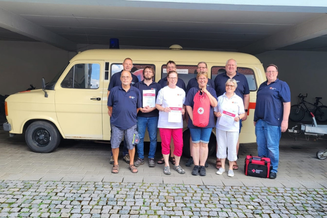 Ein Gruppenfoto der Teilnehmer vom Erste Hilfe mit Selbstschutzinhalten Modul 3. Sie stehen vor einem gelben Auto und manche halten eine Urkunde.