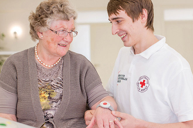 Ältere Frau mit einem Hausnotruf Armband am Handgelenk und ein junger Mann in einem Weißem Shirt mit dem Logo des Deutschen Roten Kreuz drauf